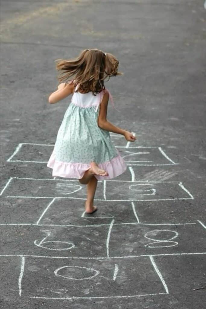 Child playing hopscotch on asphalt, wearing a summer dress, captured mid-jump with chalked numbers visible.