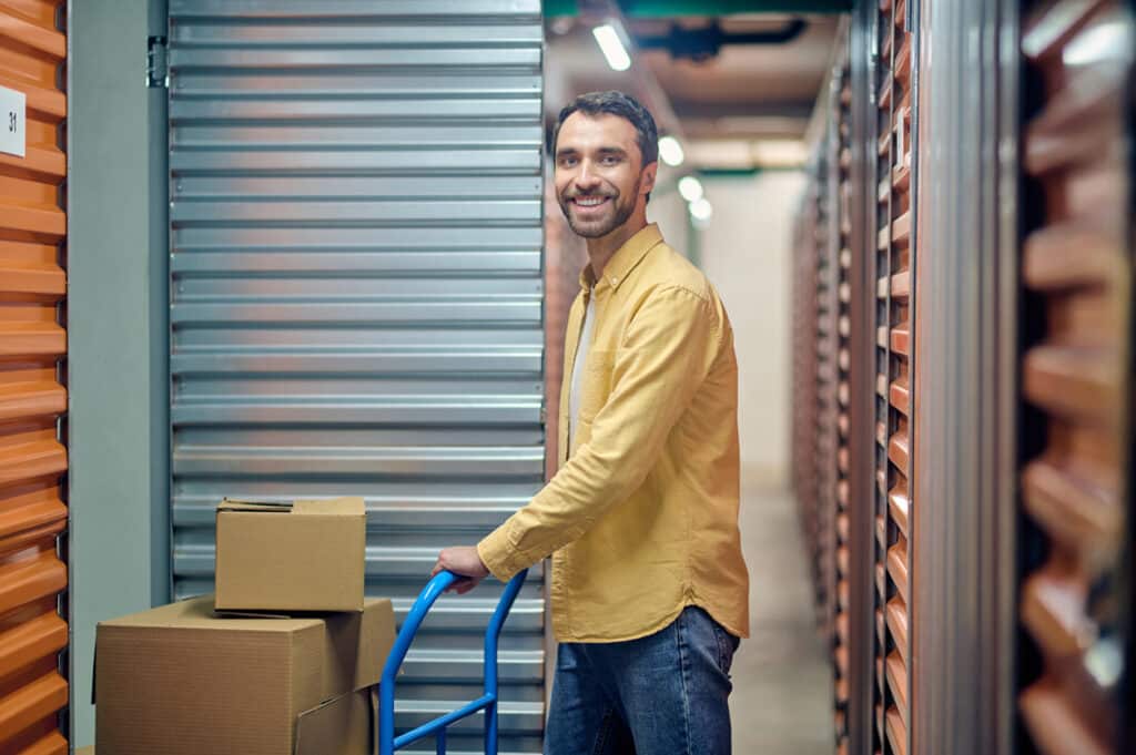 Man with cart and boxes in a storage facility aisle, smiling confidently.