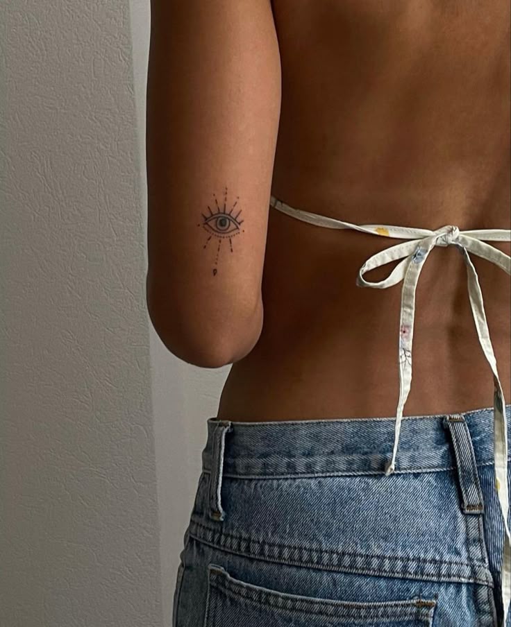 Tattooed arm with eye design, wearing a bikini top and jeans, facing a white wall background.