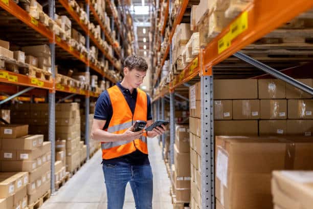 image Warehouse worker in orange vest using a tablet for inventory management among stacked shelves of boxes. | Sky Rye Design Warehouse worker in orange vest using a tablet for inventory management among stacked shelves of boxes.