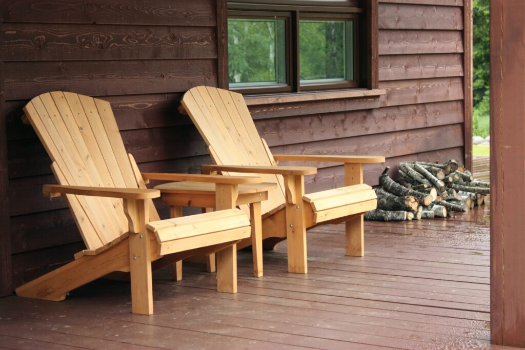 Wooden Adirondack chairs on a cabin porch with stacked firewood nearby.