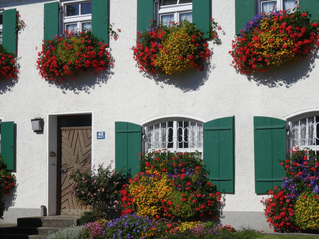 White house with green shutters and vibrant flower baskets in front, showcasing colorful blooms and a wooden door.