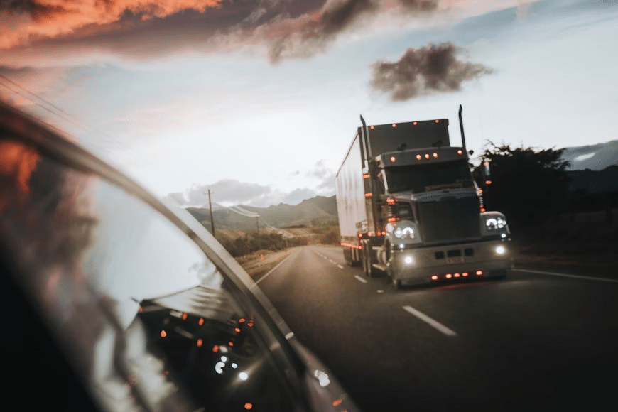 Semi-truck driving on scenic highway at sunset, reflected in car mirror.
