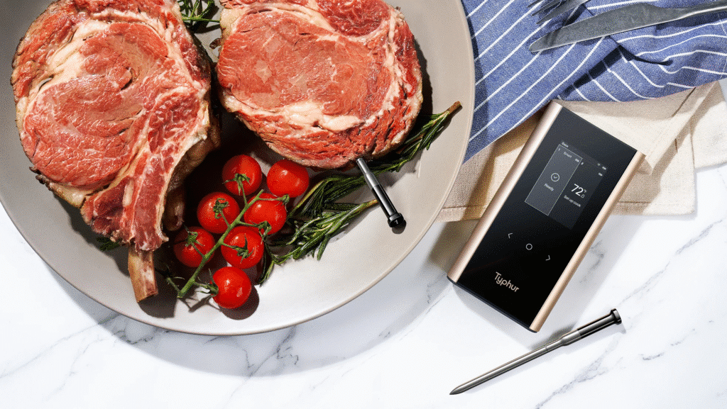 Two raw steaks with tomatoes and herbs beside a meat thermometer on a marble surface.