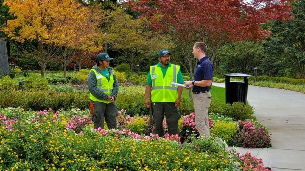 Landscapers in safety vests discuss plans in a colorful garden, surrounded by autumn foliage and vibrant flowers.