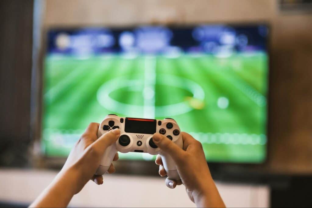 Person playing soccer video game with a white controller in front of a blurred TV screen.