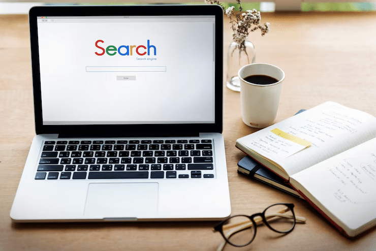 Open laptop with a search engine on screen, beside a notebook, glasses, and a coffee cup on a wooden desk.