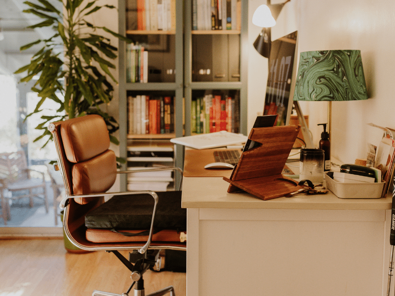 Cozy home office with leather chair, wooden desk, laptop, and bookshelves, featuring plants and calming decor.