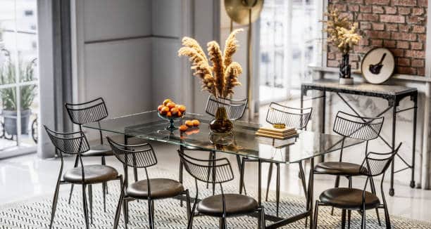 Modern dining room with glass table, black wire chairs, pampas grass centerpiece, and fruit bowl in elegant setting.