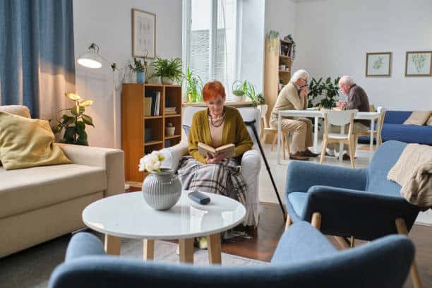 Elderly woman reading book in cozy common area of a care home; two people chatting at a table in the background.