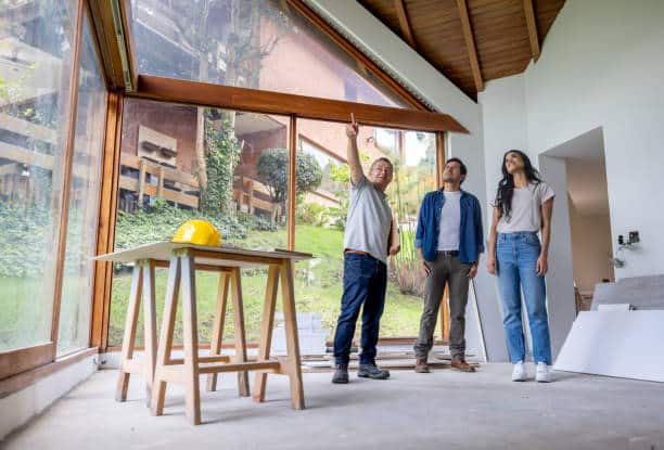 Architect showing renovation plans to a couple inside a house with large windows and a construction helmet on a table.