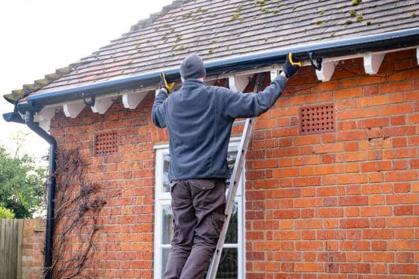 Man on ladder cleaning gutters of a brick house wearing a beanie and gloves, maintaining home exterior.