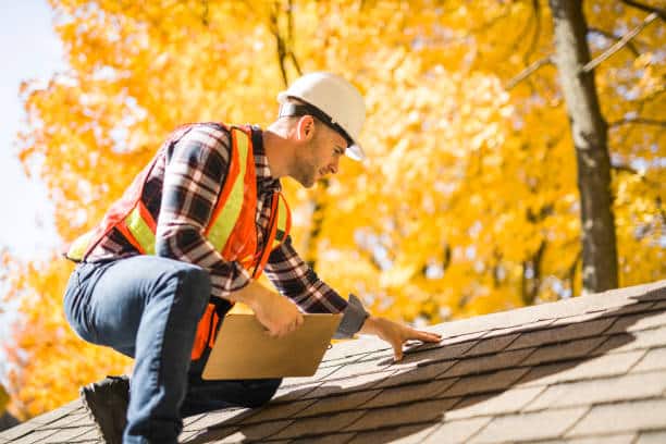 A man with hard hat standing on steps inspecting house roof Worker inspecting roof shingles in autumn, wearing safety gear and helmet. | Sky Rye Design Worker inspecting roof shingles in autumn, wearing safety gear and helmet.