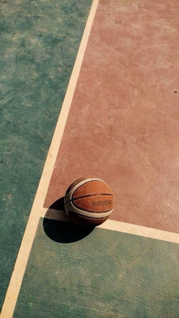 Basketball lying on a colorful court under sunlight, positioned near intersecting lines.