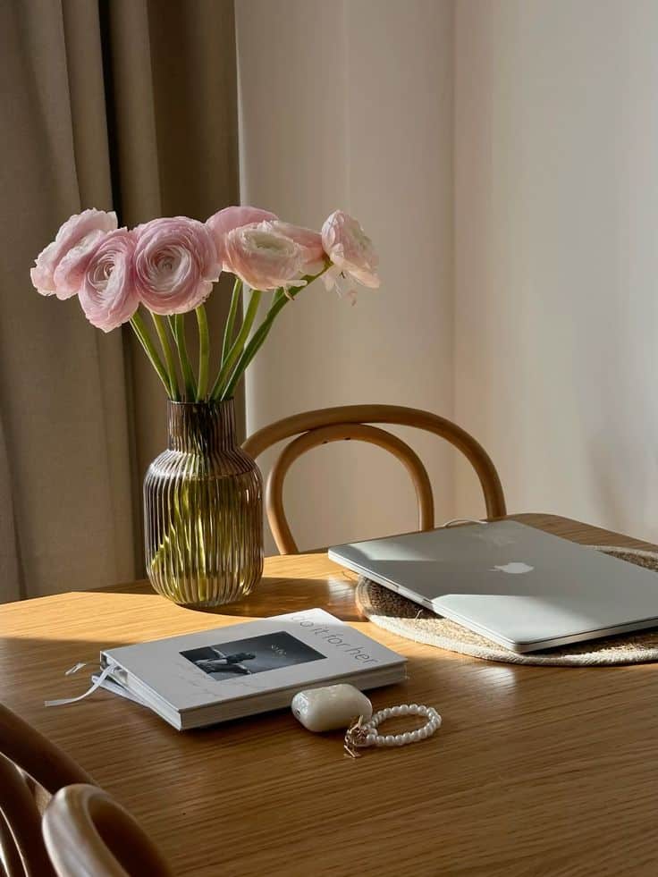 Cozy workspace with a laptop, book, and pink flowers in a vase on a wooden table, bathed in sunlight.