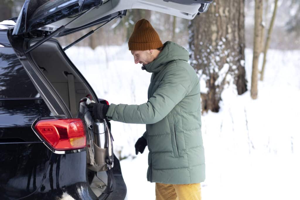 Man in winter attire packing thermos into car during snowy outdoor adventure.