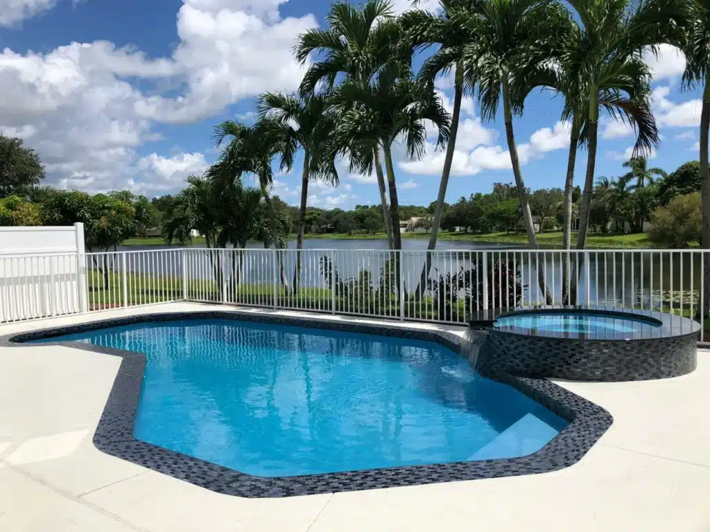 Backyard pool with a spa, surrounded by palm trees, overlooking a serene lake under a partly cloudy sky.