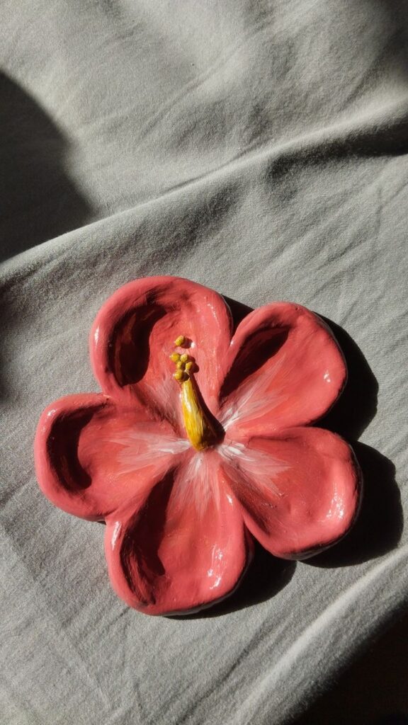 Clay hibiscus flower sculpture with pink petals and yellow stamen on a soft gray background, illuminated by sunlight.