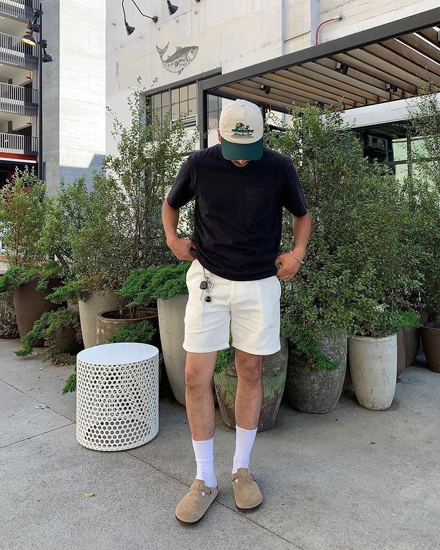 Casual outfit with black shirt, white shorts, and cap in urban setting near potted plants and modern furniture.