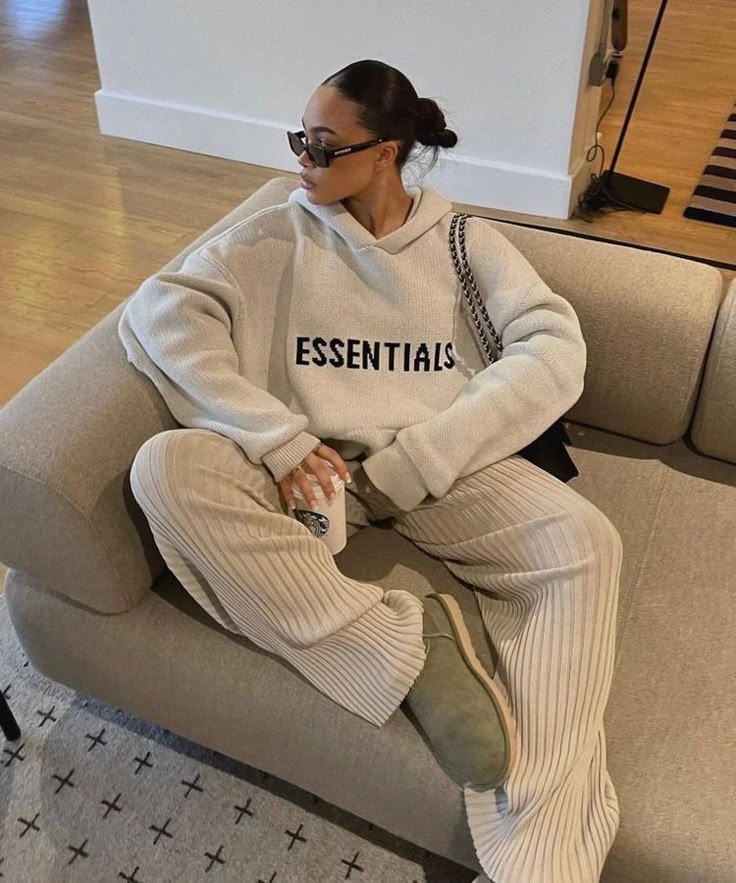 Trendy woman in cozy Essentials sweatshirt and ribbed pants lounging on sofa with coffee, sunglasses on.