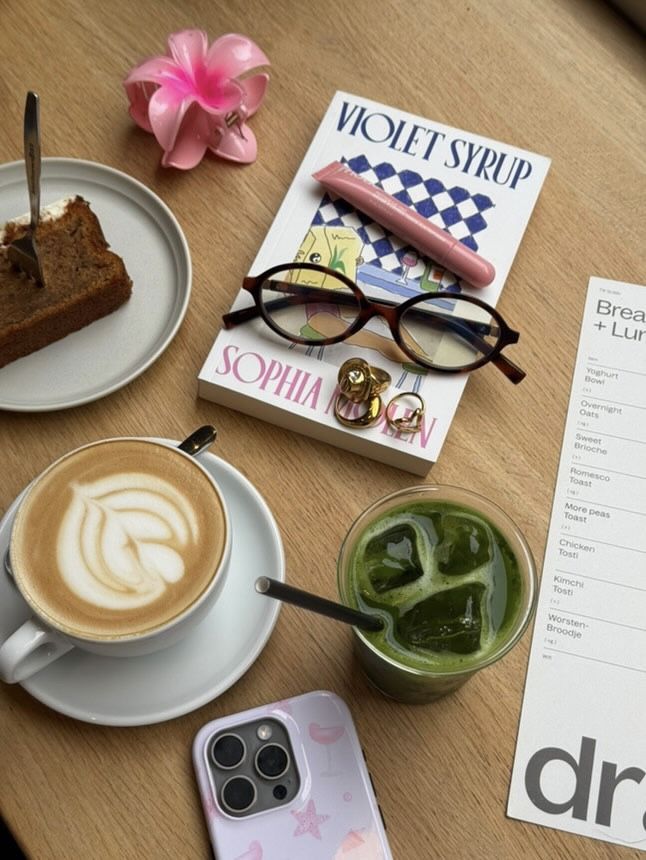 Cafe table with a latte, green juice, cake, glasses, book, pink flower, and phone on a wooden surface.