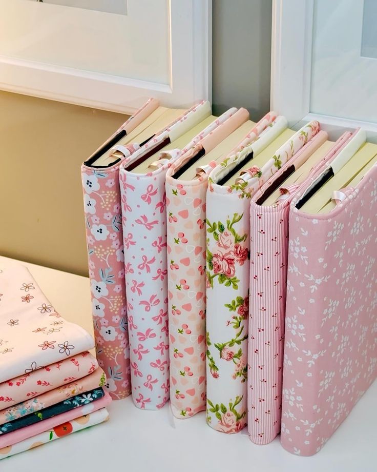 Pastel floral and patterned fabric-covered books neatly arranged on a shelf, next to a stack of folded fabrics.