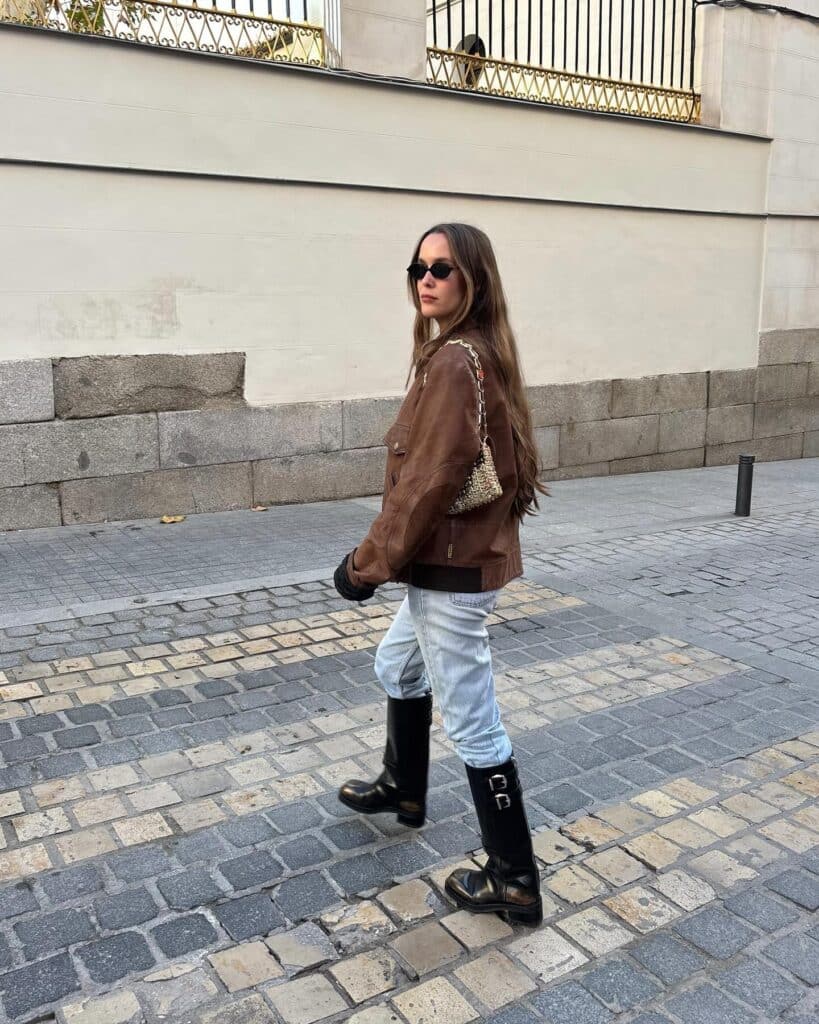 Woman in brown jacket and boots walking on cobblestone street, stylish outdoor fashion.