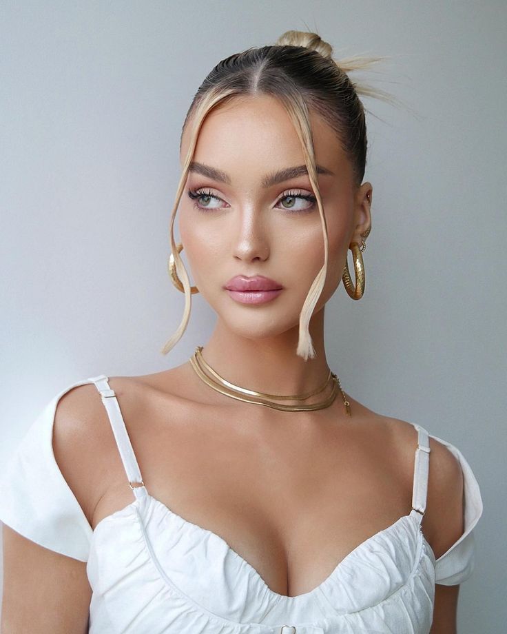 Woman with stylish updo, wearing gold hoop earrings and layered necklaces, in a white top, looking to the side.