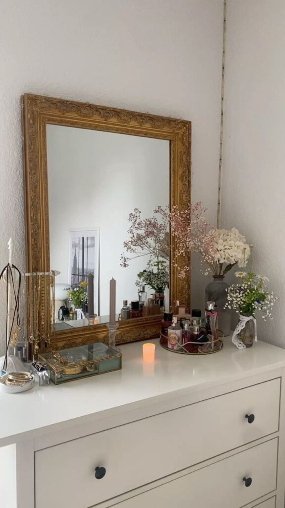 Elegant vanity setup with a mirror, flowers, perfumes, and jewelry on a white dresser. Cozy and stylish decor.