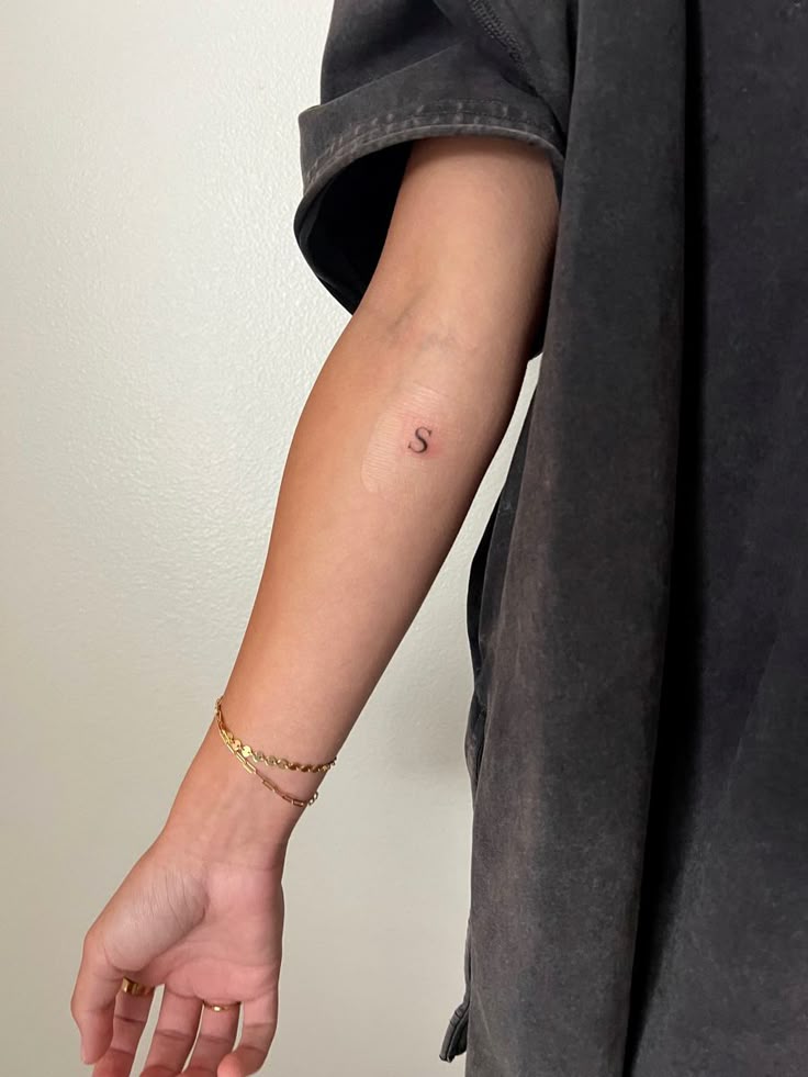 Arm with a small letter S tattoo near the elbow, wearing a black shirt and gold bracelet.