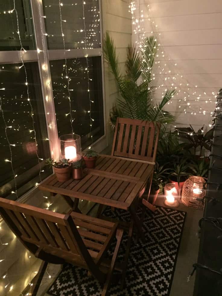 Cozy balcony with string lights, wooden furniture, candles, plants, and a patterned rug at night.