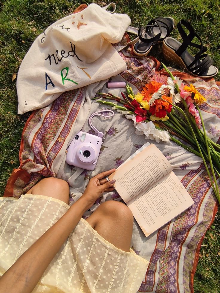 Woman enjoying a sunny picnic with a book, flowers, camera, sandals, and a need art tote on a colorful blanket.
