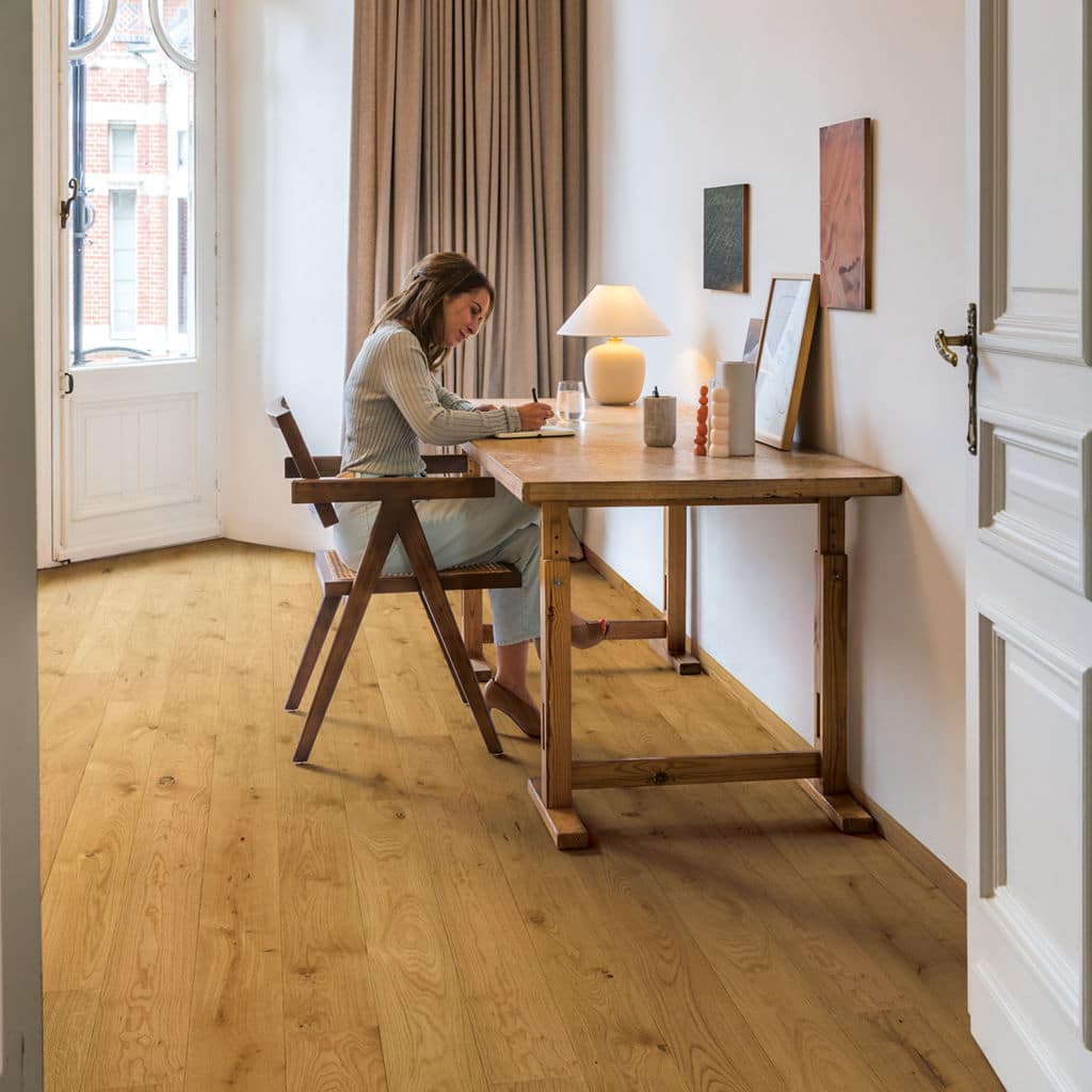 Woman writing at a wooden desk in a minimalist home office with natural light and wooden flooring.
