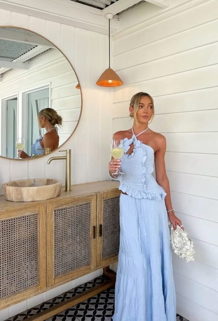 Elegant woman in a blue dress holding a drink, standing by a wooden vanity with a mirror in a stylish setting.
