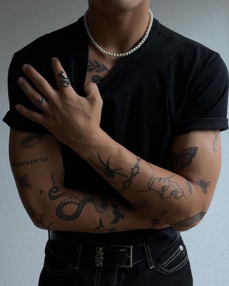 Man in black shirt, arm tattoos, wearing silver rings and pearl necklace, stands with arms crossed.