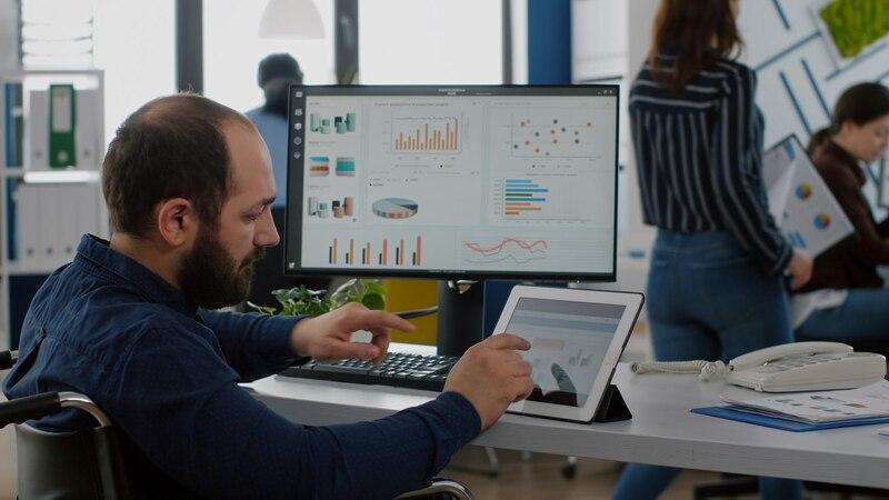 Man analyzing data on a tablet and computer in an office setting with charts and graphs on screens.