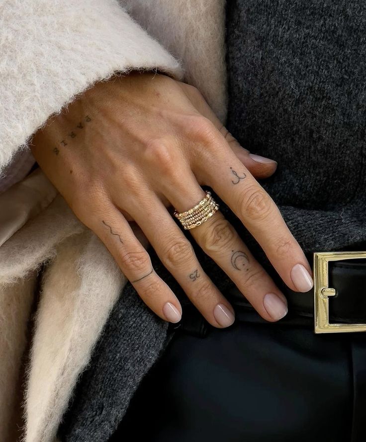 _ Hand with minimalist tattoos and stacked gold rings, wearing cozy winter coat and dark pants, close-up. | Sky Rye Design Hand with minimalist tattoos and stacked gold rings, wearing cozy winter coat and dark pants, close-up.