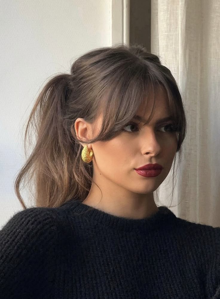 _ Woman with long hair in a ponytail, wearing a black sweater and gold hoop earrings, looking pensive near a window. | Sky Rye Design Woman with long hair in a ponytail, wearing a black sweater and gold hoop earrings, looking pensive near a window.