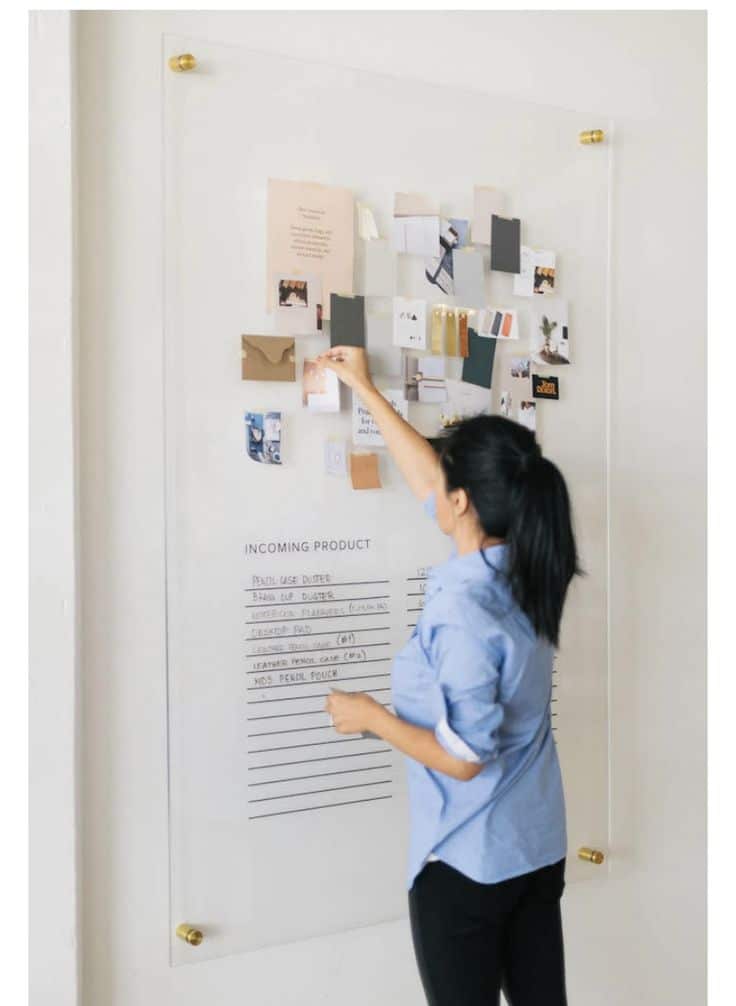 Person organizing a wall-mounted clear board with notes and product lists, dressed in a blue shirt.