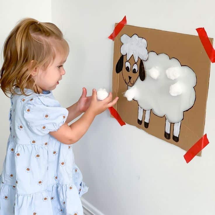 Child playing with cotton ball sheep craft on cardboard, taped to wall, wearing blue dress.