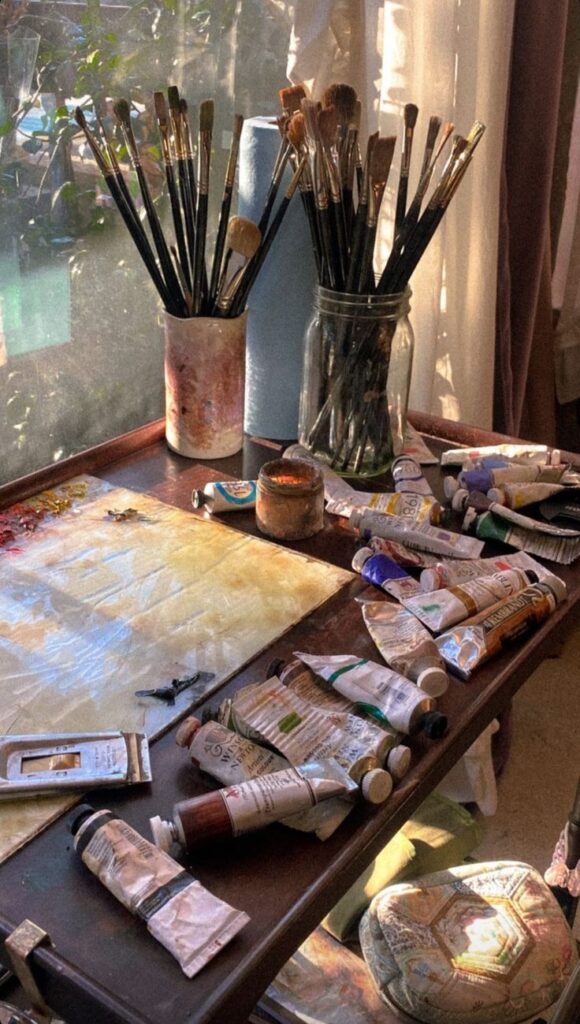 _ Artist's workspace with brushes, paint tubes, and supplies on a sunlit wooden table near a window. | Sky Rye Design Artist's workspace with brushes, paint tubes, and supplies on a sunlit wooden table near a window.