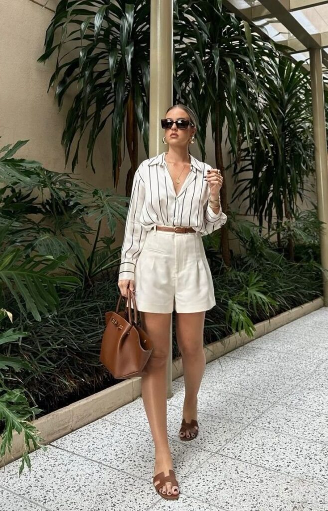 Fashionable woman in striped blouse and white shorts, carrying a brown handbag, walking in garden atrium.