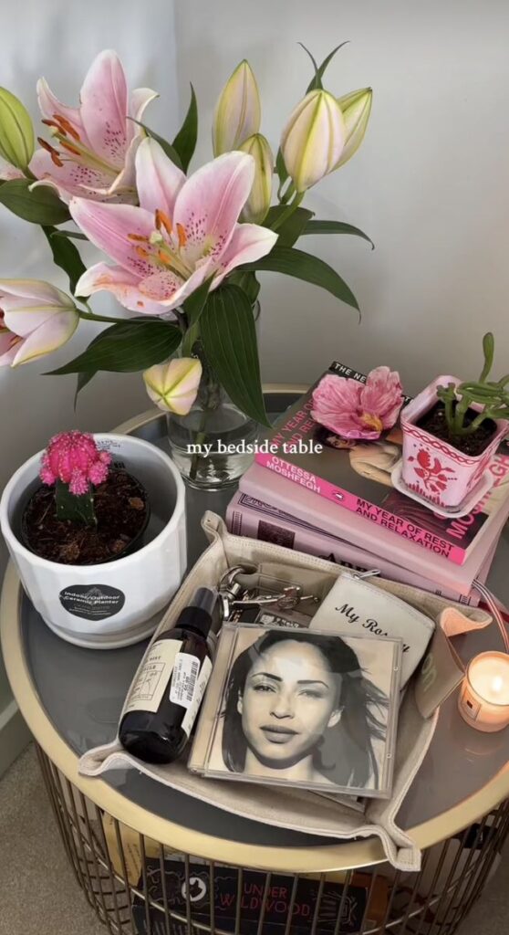Bedside table with pink lilies, books, candle, potted cactus, and skincare essentials for a cozy, aesthetic setup.