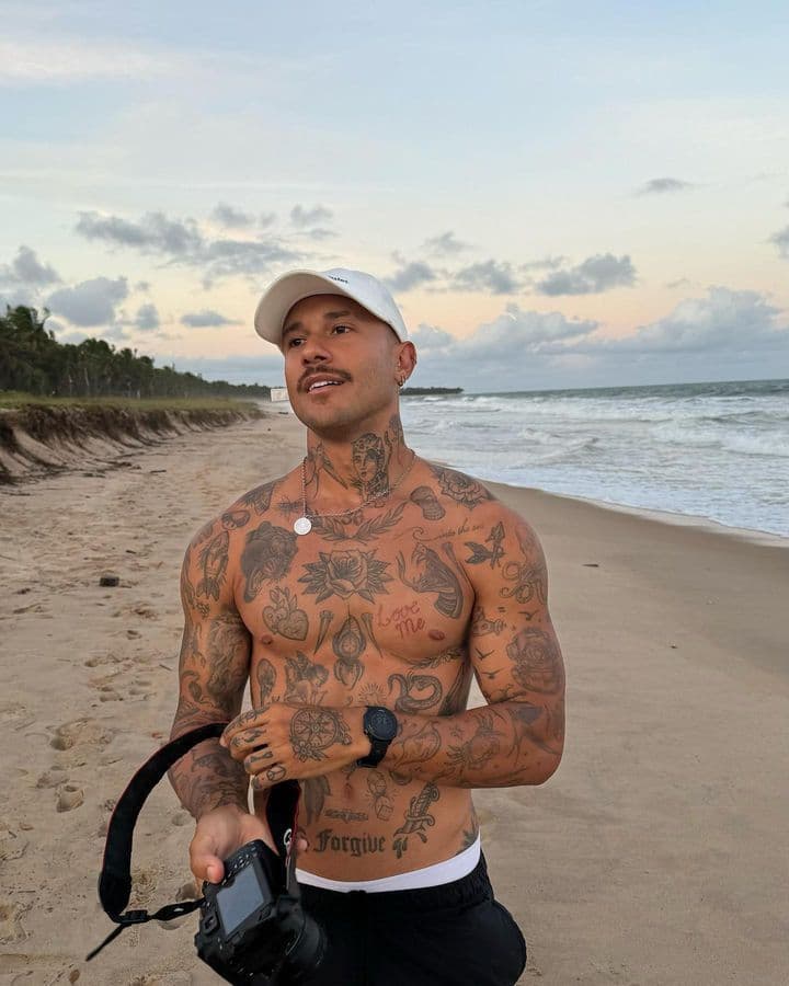 Tattooed man holding a camera on a serene beach at sunset, wearing a white cap and black pants.
