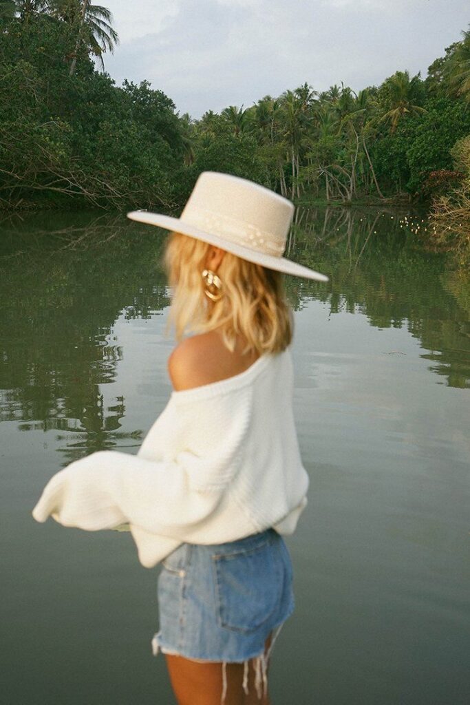 Woman in white sweater and hat by a serene lake surrounded by lush greenery.