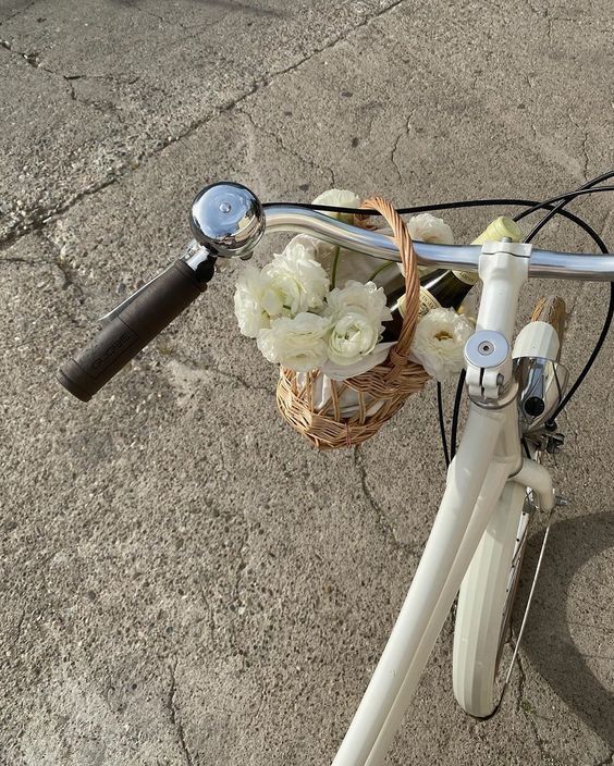White bicycle with wicker basket holding white flowers on a sunny day, parked on pavement.