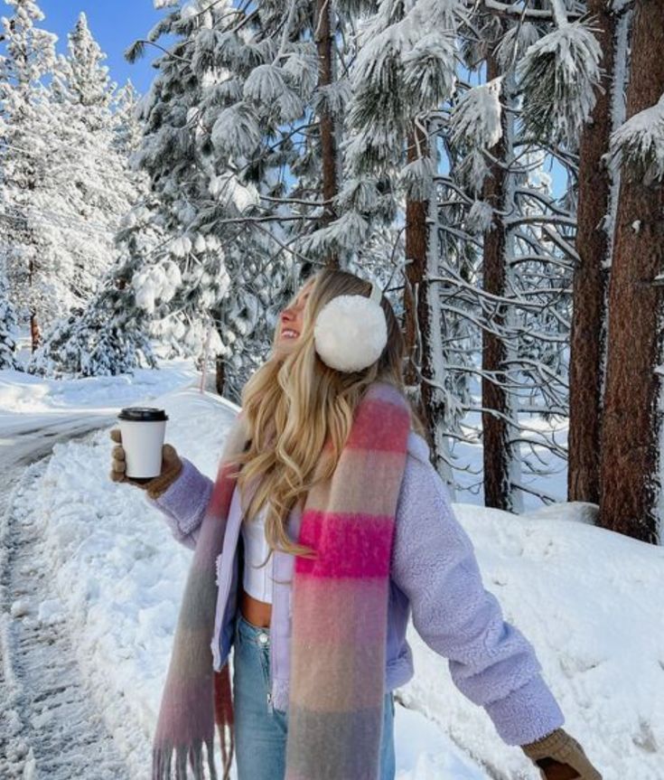 Woman enjoys snowy forest with hot drink, wearing earmuffs, colorful scarf, and cozy jacket.