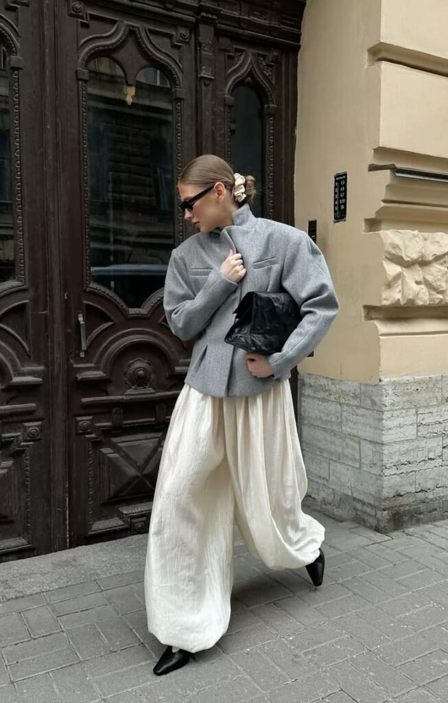 Fashionable person in gray jacket and white trousers, carrying a black clutch, walking past an ornate building door.