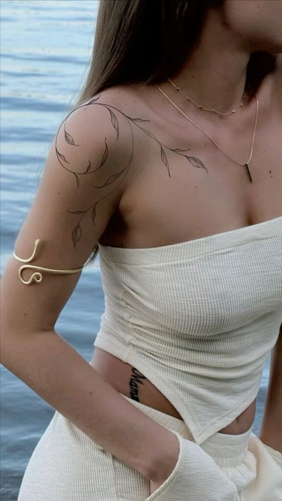 Woman in a strapless top by water, wearing a gold arm cuff and leaf tattoo on shoulder.