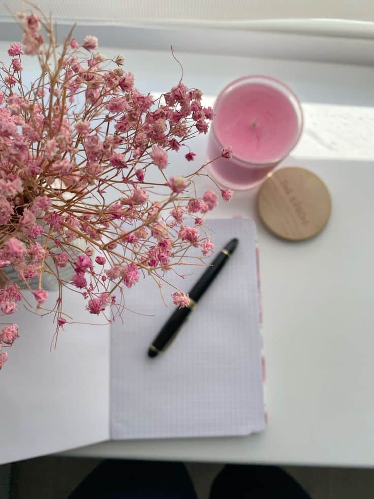 Open notebook with pen, pink candle, and dried flowers on a white table, creating a cozy workspace ambiance.